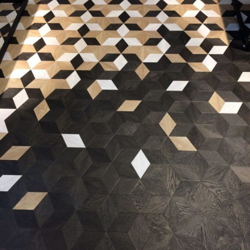 Close-up of intricate floor tiling with a geometric, three-dimensional cube pattern in black, white, and tan wood-look tiles. The perspective is from above, showcasing the detailed arrangement of the floor tiling and the play of light and shadow.
