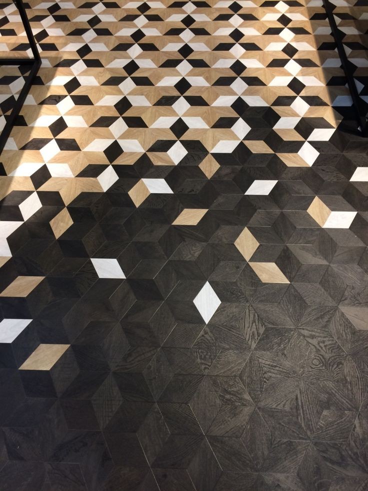 Close-up of intricate floor tiling with a geometric, three-dimensional cube pattern in black, white, and tan wood-look tiles. The perspective is from above, showcasing the detailed arrangement of the floor tiling and the play of light and shadow.