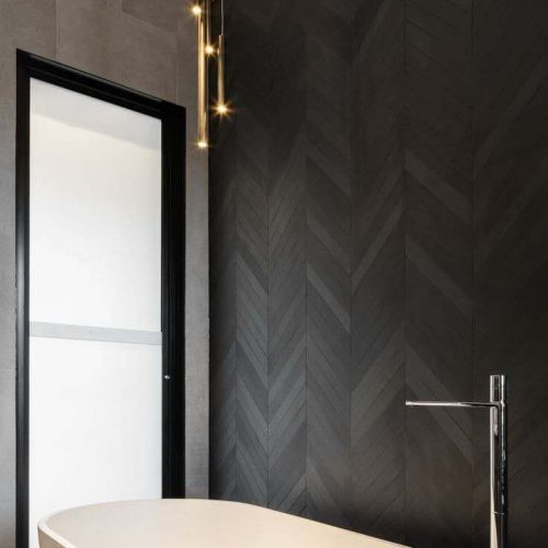 A modern bathroom featuring a freestanding white oval bathtub. The wall behind the tub is adorned with dark, possibly charcoal or black, chevron-patterned bathroom tiling, while the adjacent wall and floor have large format grey tiles.
