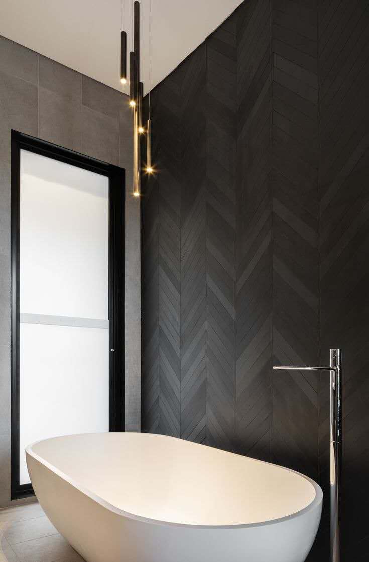 A modern bathroom featuring a freestanding white oval bathtub. The wall behind the tub is adorned with dark, possibly charcoal or black, chevron-patterned bathroom tiling, while the adjacent wall and floor have large format grey tiles.